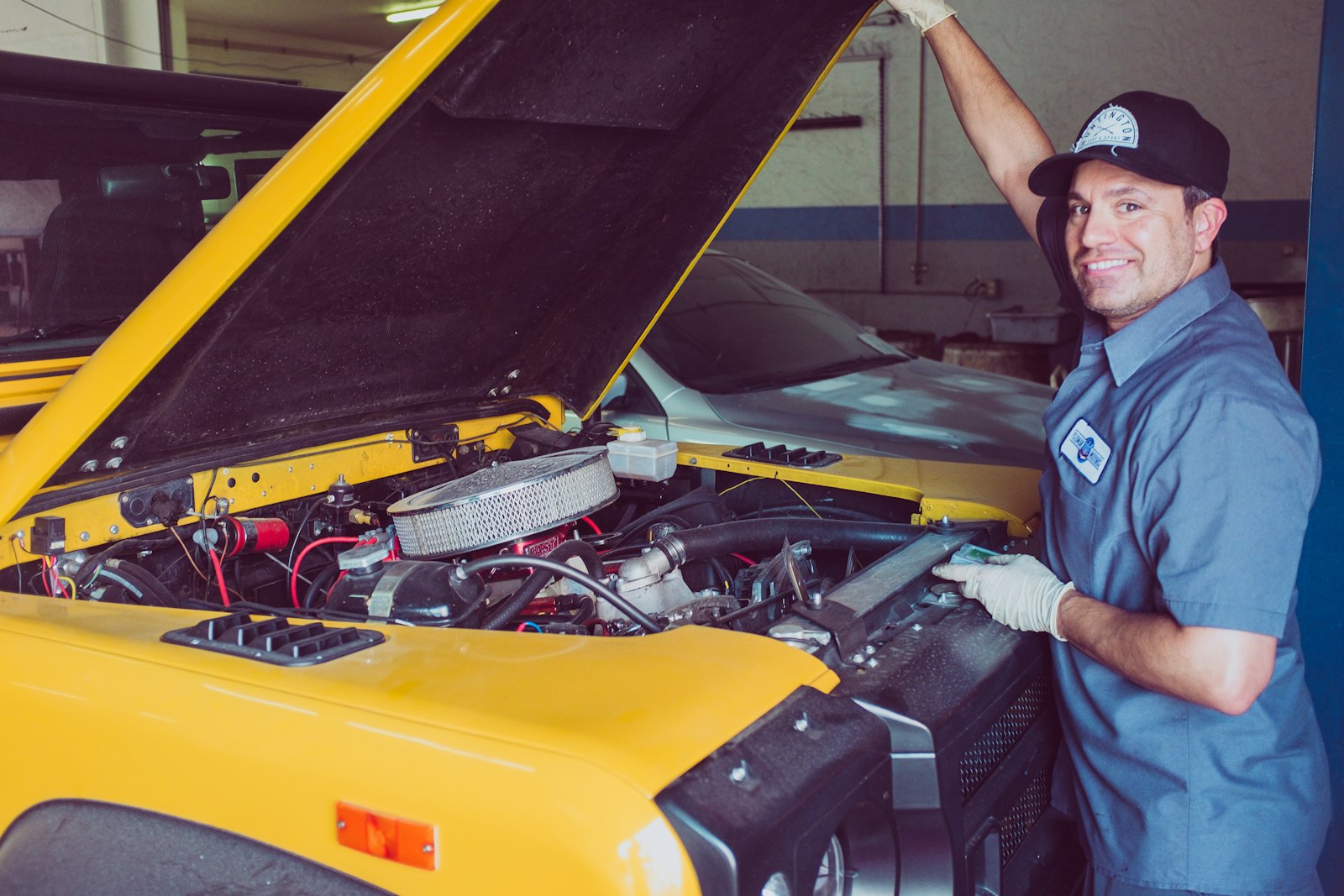A smiling Marion Auto Works technician in blue coveralls beside a bright yellow car with its hood raised